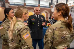 A man in a decorated military dress uniform talks and gestures with three women in camouflage uniforms inside a large.
