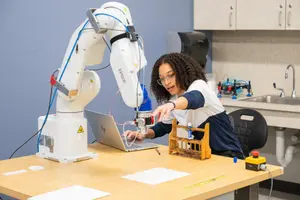 A woman with curly hair reaches toward a wooden test tube rack while sitting at a table with a robotic arm and laptop.