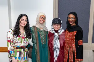 Four women stand side by side smiling, wearing colorful traditional and modern clothing with headscarves and accessories.