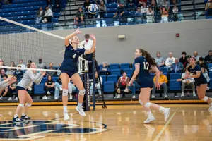 A volleyball player in a navy uniform jumps to set the ball while teammates watch and prepare on the court.