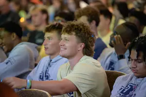 A group of young men sit closely together, some smiling and engaged, while one looks down with a serious expression.